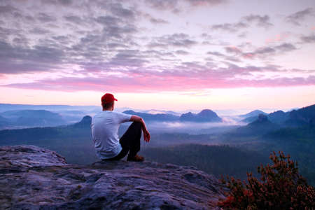 Adult sportsman in white shirt, dark trousars and red cap. Man sit on sharp cliff above valley in rocky mountains park and watch into landscapeの写真素材