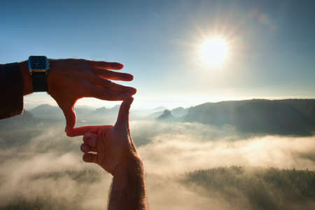 Close up of hands making frame gesture. Blue misty valley bellow rocky peak. Sunny spring daybreak in mountains.の写真素材
