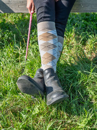 Crossed legs wear riding paddock boots and long knee socks. Horse rider hold wisp while sit in paddock of horse riding club.の写真素材