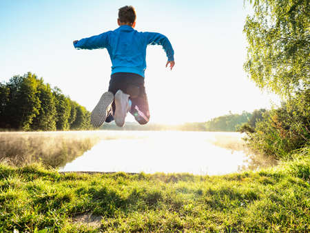 Crazy boy is making fun and jumping on lake bank against evening sunset. Funny childhood.