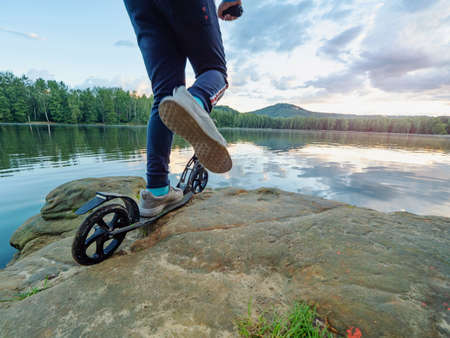 Blue sports wearing boy stay with fun push scooter on cliff at lake and makes tricks. Evening sun at horizon makes flares.の写真素材