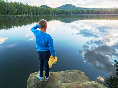 Blue wearing boy with yellow towel and swimming goggles in hands stay on jumping cliff above lake water. Natural summer holiday resort.の写真素材