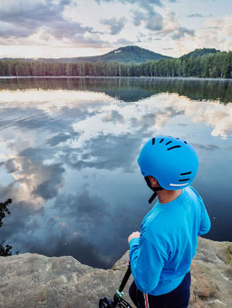 Cute boy with blue skater helmet arrived by push scooter to lake. Summer sunset in natural park.の写真素材