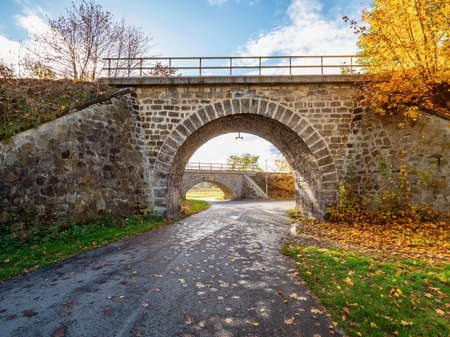 Golden leaves on trees. Old stony viaduct wiith railways over thin local asphalt road.