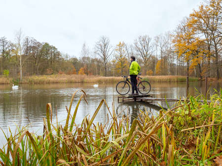Cyclist stay at his trekking bike on small pond pier and watching swans and opposite bank.の写真素材