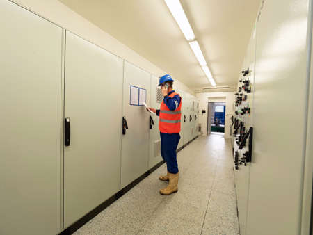 Expert in reflective vest stay in front of switch board, reading data from document and call...