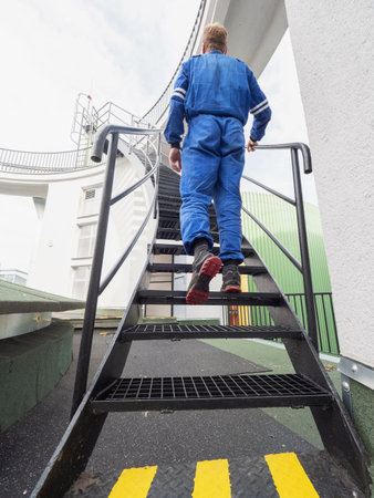 Manipulator in chemical operation wearing a blue overall and walks up a steel spiral staircase to the upper floor.の写真素材