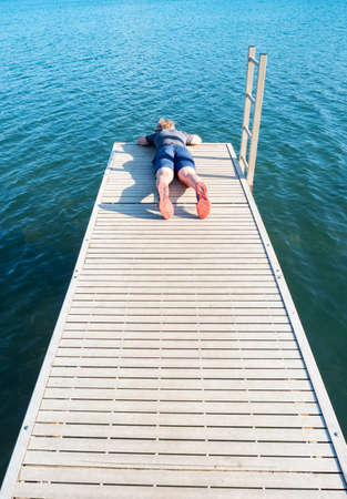 Young blond boy lying on a wooden pier over a lake watch fish in blue water below. The boy enjoys his childhood.の写真素材