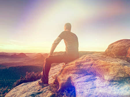 Tourist man sit on a stone at cliff edge overlooking the plains below at misty border of horizon. Rear view. Abstract lighting, colorful flare.の写真素材