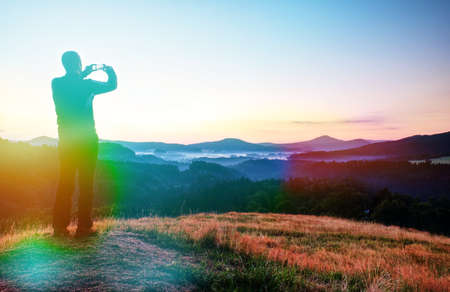 Man hold phone above head and takes photos of morning misty landscape. Abstract lighting, colorful flare.の写真素材