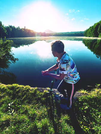 Blond hair boy with scooter stands on a park path along the lake. Childhood sports concept. Evening sun is making flares and reflections in water.の写真素材