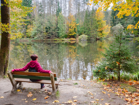 Woman in pink outdoor jacket sitting at forest pond in autumn colors. Relax at the pond.の写真素材
