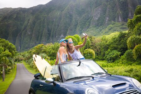 Car Road Trip vacation young people taking selfie photo with phone during summer travel vacation. Tourists couple taking photos on Hawaii in convertible car, with smartphone cameraの写真素材