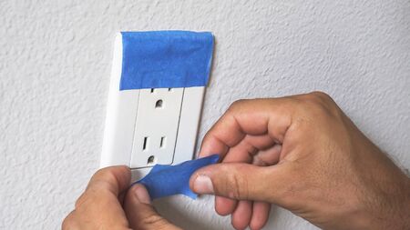 Painter Man Using Masking Blue Tape to Secure Electric Outlet. Preparation For Room Painting.