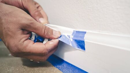 Removing masking tape from molding. A painter pulls of blue painter's tape from the wall to reveal a clean edge baseboard.の写真素材