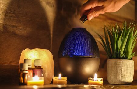 Woman adding essential oil to electric diffuser lamp, candles on wooden table in room. Aromatherapy.