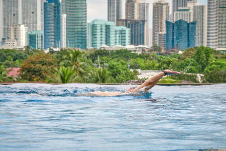 Manila, Philippines - Feb 02, 2020. A man swims a backstroke in a pool on the roof of a luxury hotel. View of the city of Manila from the pool of the luxury five-star Discovery Primea hotel. Sunny weather. Skyscrapers on the background.のeditorial素材