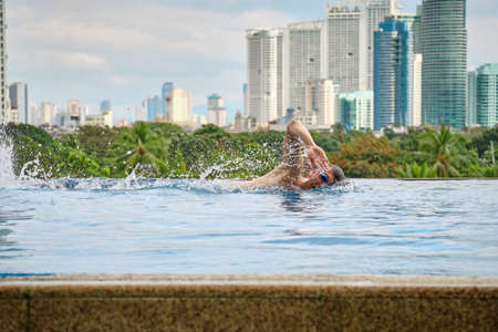 A man swims a butterfly in a pool on the roof of a luxury hotel. View of the city of Manila from the pool of the luxury five-star Discovery Primea hotel. Sunny weather. Skyscrapers on the background.のeditorial素材