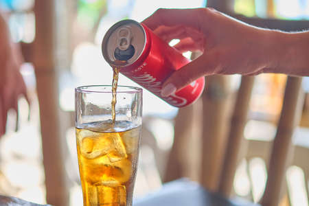 A glass of rum and coke next to a red iron can of Coca Cola against the background of a sandy beach.