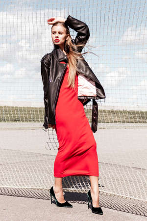 A fashionable girl with red hair in a black leather jacket poses in a fashion shoot. The wind blows a red scarf. Volleyball net.のeditorial素材