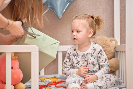 A little girl in pajamas receives birthday gifts while sitting in a white baby cot. Balloons in the background.の写真素材