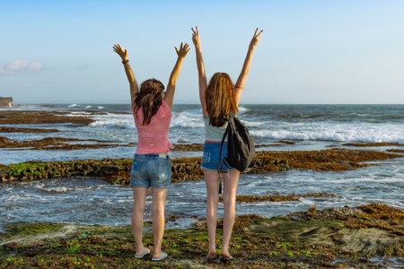 Two slender young female travelers with their backs to each other. Watch the sea towards sunset near...