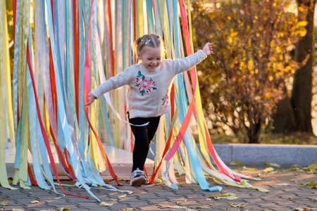 A cute little girl with pigtails in a gray sweater in an autumn park runs through colorful ribbons.の写真素材