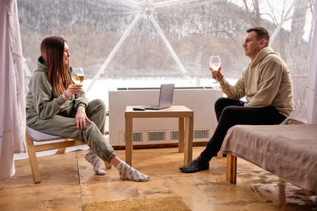 A guy and a girl with glasses of white wine look out the window in glamping. In winter, outdoors in a tent. The computer is on the table.の写真素材