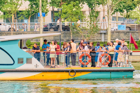 BEIJING, Tianjin - JUN 01, 2025: Modern river ferry with panoramic windows sailing past green trees and glass skyscrapers