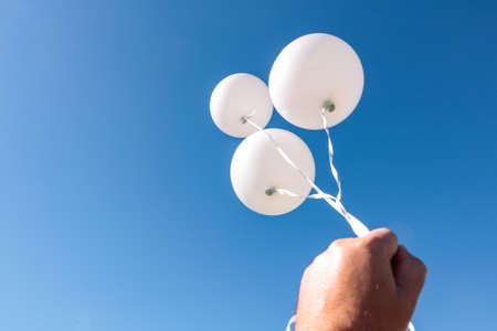 A male hand holds three white air balloons against a very blue sky.の写真素材