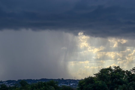 Image of rain on the horizon, with the left side showing rain and the right side showing orange clouds that apparently do not have rain. The base of the image has some trees and a city in the background, with a dark cloud above.の写真素材