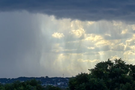 Image of rain on the horizon, with the left side showing rain and the right side showing orange clouds that apparently do not have rain. The base of the image has some trees and a city in the background, with a dark cloud above.の写真素材