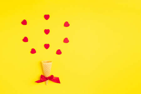 A simple but modern ice cream cone decorated with red ribbon and with red hearts flying above it on a pastel yellow background. Flat layout.の写真素材