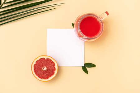 A glass jug with refreshing juice and a slice of fresh red grapefruit with a plain white paper next to a green palm leaf on a pastel sand color background. Flat layout concept.の写真素材