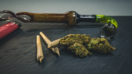 Marijuana buds and joint lie on a dark gray background. Smoking pipe and lighter near cannabis....