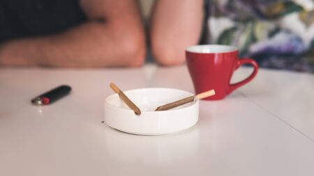 Two medical marijuana joins are in the ashtray, a cup of coffee and lighter near. Smoking marijuana joint indoors. Cannabis is a concept of herbal medicineの写真素材