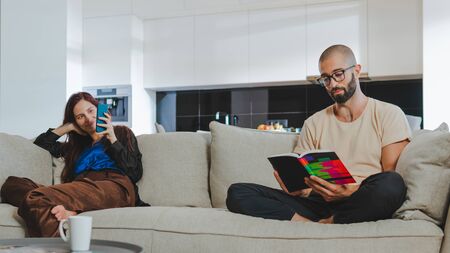 Millennial couple in love relaxing together on sofa at home. Woman using her smartphone and man reading bookの写真素材