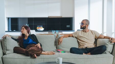 Millennial couple in love relaxing together on sofa at home. Woman using her smartphone and man reading bookの写真素材