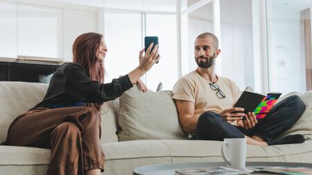 Millennial couple in love relaxing together on sofa at home. Woman using her smartphone and man reading bookの写真素材