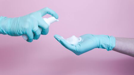 Close-up of female hands with rubber glove pumping cleansing foam on pink background. Concept of...