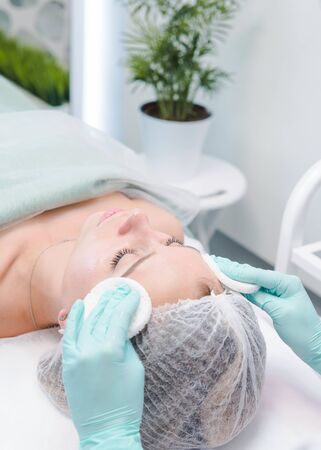 The female dermatologist cleaning face skin by cotton sponge to beautiful girl. Cosmetologist removing facial mask from female face. Concept of skin and health careの写真素材
