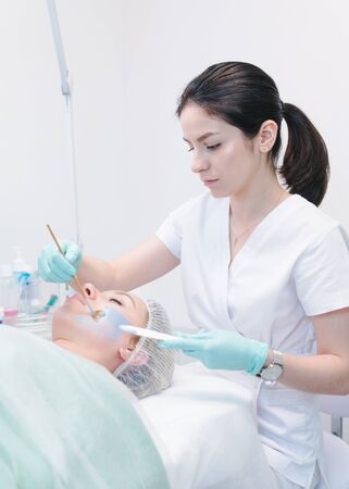 The female cosmetologist applying blue cosmetology mask to young woman face, close-up. Facial cosmetic treatment in beauty clinic. Concept of skin and health careの写真素材