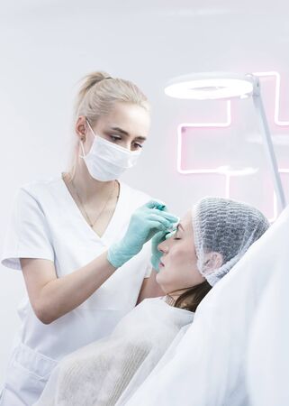 The young beautician doctor preparing to making injection in female forehead. The doctor cosmetologist makes anti-aging treatment and face lift procedure. Concept of beauty salon and plastic surgery clinic.の写真素材