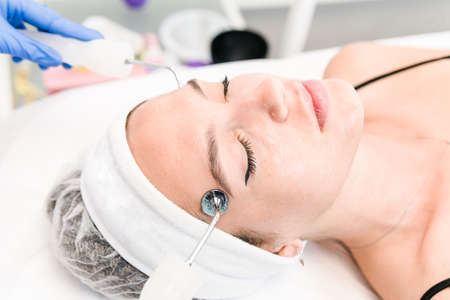 The young female client of cosmetic salon having microcurrent procedure on her face with special...
