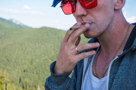 Close-up of young man smoking marijuana joint outdoors. Hemp blunt in the male hand. Cannabis is a concept of herbal and alternative medicineの写真素材