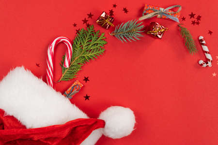 Christmas background, flat lay. Santa Claus hat with decoration and fir tree branches on red...