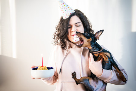 Happy young girl giving homemade cake to her dog, indoors. Concept of celebrating dog's birthday partyの写真素材