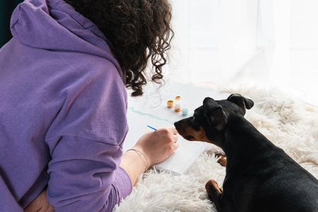 The adult woman painting by numbers indoors, her pet miniature pincher near. People with pets. Concepts of leisure activity, hobby and relaxの写真素材