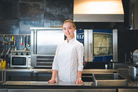 Portrait of confident and smiling young woman chef dressed in white uniform, professional kitchen are on background. restaurant kitchenの写真素材