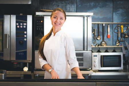 Portrait of confident and smiling young woman chef dressed in white uniform, professional kitchen...